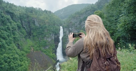 Woman taking photograph of waterfall with smartphone wearing yellow jacket photographing scenic landscape nature background view enjoying vacation travel adventure Norway
