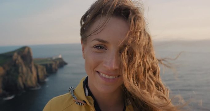 Close up portrait of Young Woman smiling with Red hair blowing in wind looking at sunset over ocean Girl wearing yellow raincoat trekking in Scotland Slow Motion