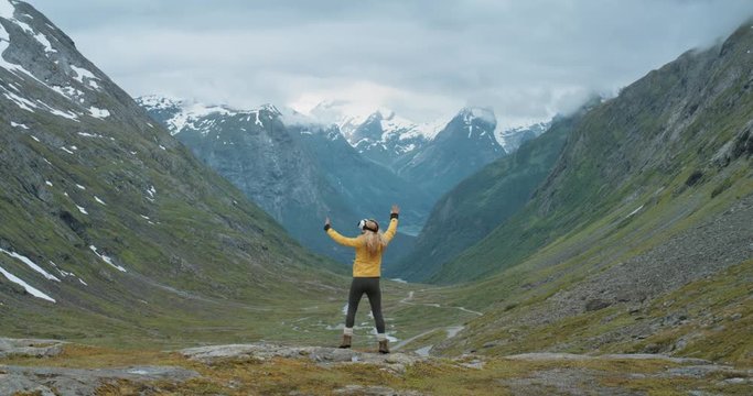 Brave woman traveler in Norway wearing virtual reality headset looking at mountain view enjoying outdoor travel experience  watching 360 video imagination concept wearing yellow jacket