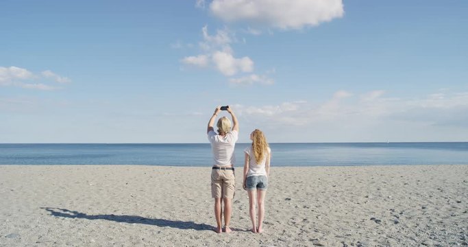 Young couple looking towards ocean taking photo smart phone enjoying summer fun vacation travel concept