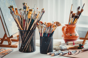 Artistic equipment: easel,  paintbrushes, tubes of paint, palette and paintings on work table in a artist studio.