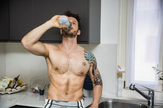 Front view of sexy muscular topless man in kitchen at home, eating an apple, wearing only boxer shorts, showing big muscles