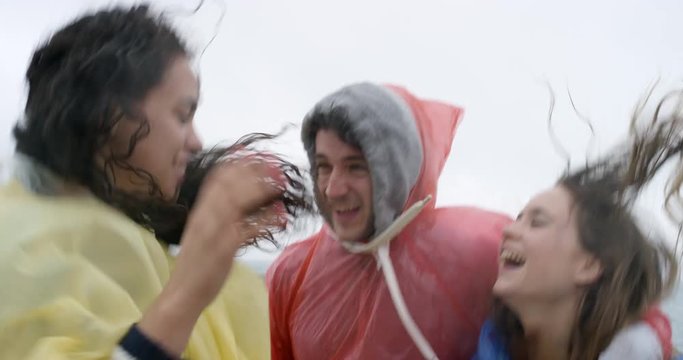 Diverse group of friends wearing multi colored poncho rain coat dancing freestyle silly dance in rainy weather on the beach celebrating adventure