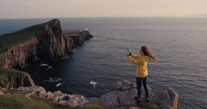 Adventure Woman taking photograph of sunset ocean view with smartphone photographing scenic landscape nature background enjoying vacation travel. Neist Point Lighthouse, Isle of Skye, Scotland