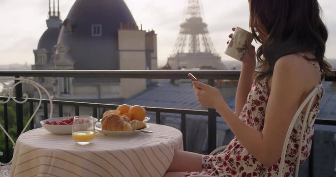 Beautiful young woman holding smartphone digital touchscreen sitting on Hotel Balcony enjoying sunrise view of Paris skyline Eiffel Tower in Background