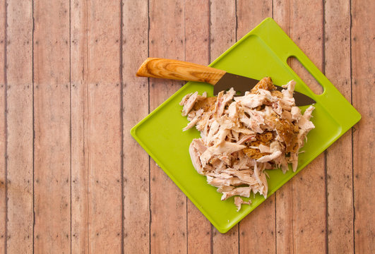 Shredded rotisserie chicken on a green cutting board and carving knife against wood plank background