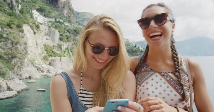 Two young women hanging out at beach using mobile phone sending text message snapchat sharing digital content on social media tourist girls enjoying summer vacation on Amalfi Coast Italy