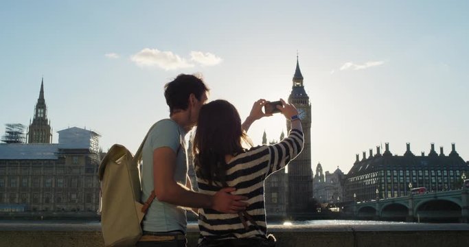 Tourist couple taking photo of Big Ben London with smart phone technology for social media at sunset enjoying vacation travel adventure