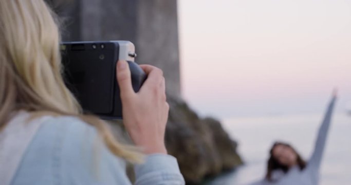 Young hipster woman taking photo of best friend with polaroid camera at sunset on beach Girl photographing sharing  summer vacation 