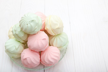 Multicolored marshmallows in a glass plate on the table. White wooden background. Top view with space for text.