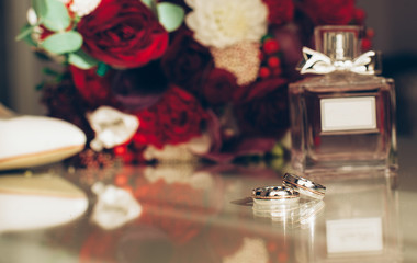 A bouquet of white and red flowers with white ribbons, beige shoes, perfume, rings, boutonniere, and a card on the table.