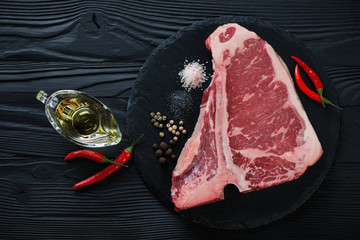 Stone slate tray with raw black angus club steak and condiments, top view over black wooden surface
