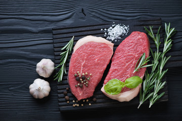 Black wooden cutting board with two raw fresh beef medallions, fresh rosemary, pepper, sea salt and garlic, top view