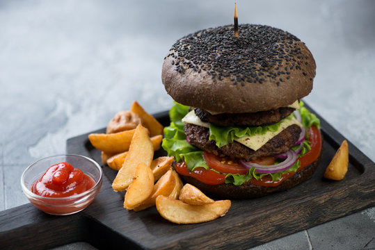 Black double beef burger on a wooden serving board with potato wedges, selective focus, shallow depth of field