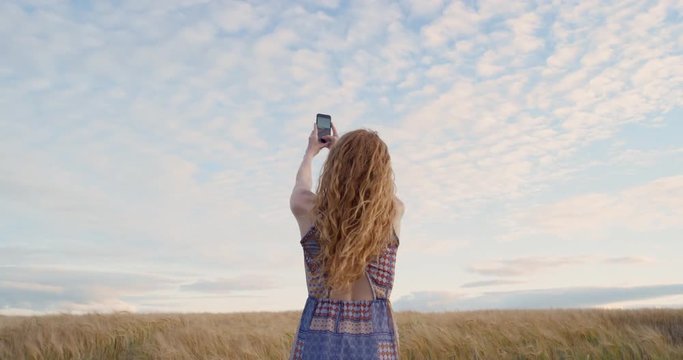 Rear view of attractive red head woman taking panorama photographing wheat field using smartphone capturing epic nature background 