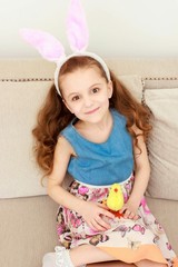 Cute little girl with bunny ears and cuddly toy sitting on sofa at home