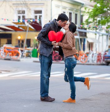 photo of cute couple holding heart shaped pillow and kissing on the wonderful street background