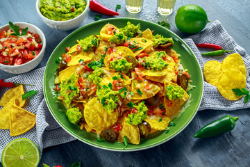 Nachos with cheese, jalapeno peppers, red onion, parsley, tomato, salsa, guacamole sauce and tequila on green plate.