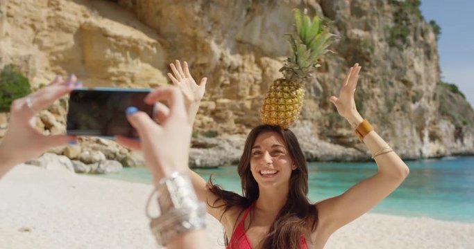 Happy teenage girl friends taking photo using mobile smart phone balancing pineapple on head posing for camera being silly on tropical beach summer vacation