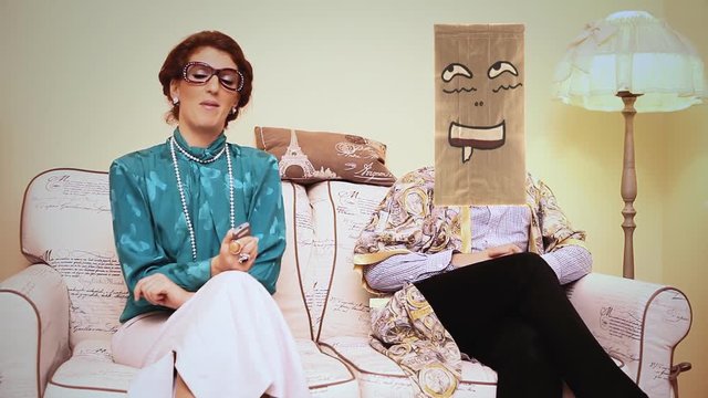 A verbose harsh wife keeps talking and watching the tv, while the husband gets excited thinking about something else (he wears a breadbag face). Funny comedic shot.
