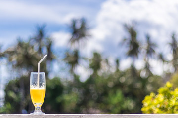Glass of orange juice on bright tropical blurred background with palms, outdoors. Tropical Bali island, Indonesia.