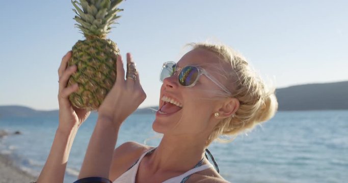 Close up attractive woman holding up pineapple posing for camera smiling laughing pretending to eat fruit at sunset on tropical beach