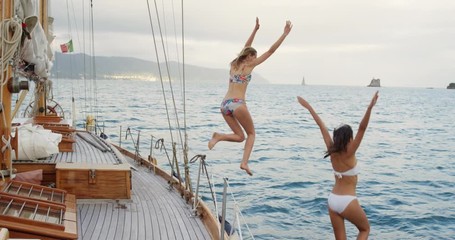 Beautiful girl friends jumping into blue sea from luxury wooden sailboat fun happy lifestyle ocean vacation travel