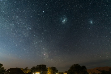 The starry sky and the majestic Magellanic Clouds, outstandingly bright, captured in Africa. Acacia trees and straw hut in the foreground. Adventure into the wild. - obrazy, fototapety, plakaty