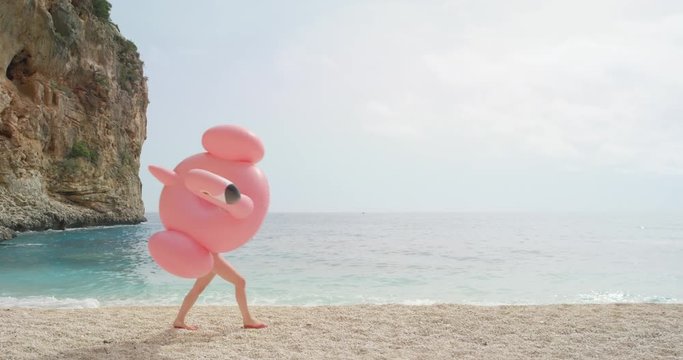 Woman walking on empty beach holding giant inflatable flamingo enjoying summer vacation on tropical beach holiday wearing full one piece swimsuit