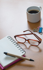 blank open notebook on a brown table with a cup of coffee and eye glasses