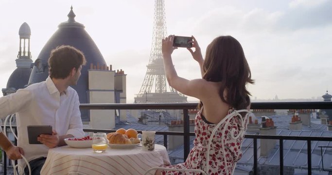 Young tourist couple in Paris hotel enjoying breakfast on terrace woman photographing view of Eiffel Tower at Sunrise with smartphone in the morning