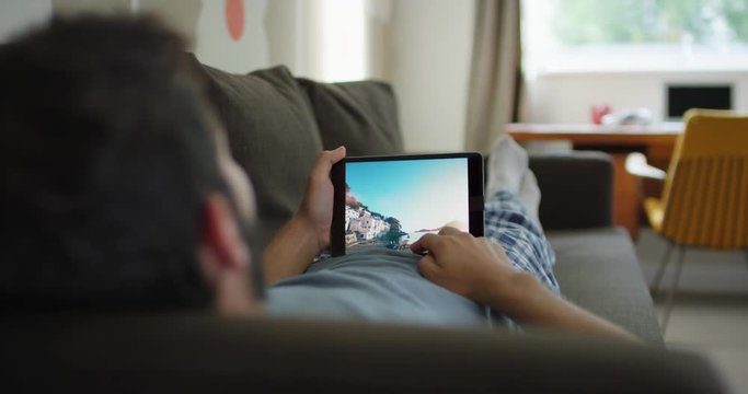 Closeup of man using smartphone at home lying on sofa viewing travel photos on touch screen browsing social media sharing wanderlust inspiration on digital device