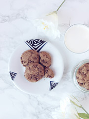 Crisp chocolate cookies on plate with a glass of milk and white flower on marble background. Flat lay. Top view