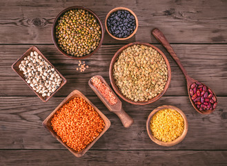 Set of various beans and lentils in bowls and scoop on wooden background, top view