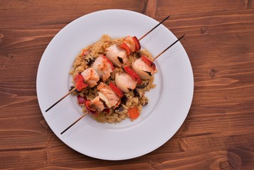 Grilled salomon skewer consist from salmon meat, onion and red sweet pepper served with wholegrain couscous mixed with vegetables, carrot, challot and aubergine server on white plate and wooden table 