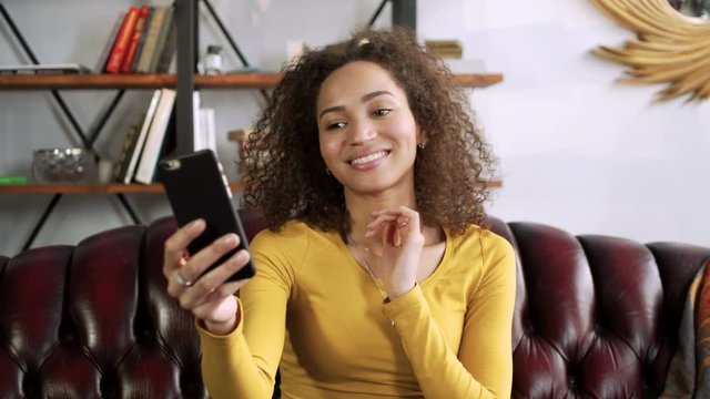 young happy latin woman smiling and laughing using mobile phone for video chat online with friends and blogging for social media while sitting in room at cozy home during sunny day