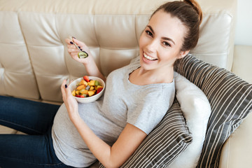 Obraz na płótnie Canvas Happy pregnant woman sitting and eating fruit salad on sofa