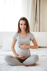 Cheerful pregnant woman sitting on bed with legs crossed
