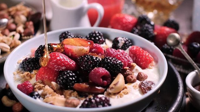 Slow motion view: pouring honey over a bowl of cereals with berries for breakfast with dry fruits in the defocused background