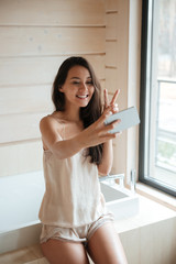 Cheerful cute young woman taking selfie with smartphone in bathroom