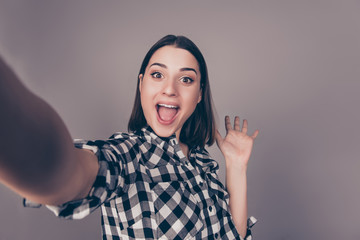 Hi, how are you doing? A young cheerful girl in checkered shirt taking a selfie, smiling in camera and waving her hand