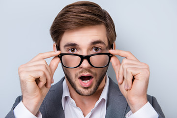 Close-up portrait of amased surprised handsome young man with bristle in formal wear in shock and holding glasses while standing on gray background