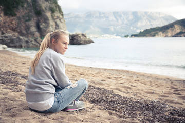 Fototapeta na wymiar Young smiling casual woman sitting on beach with rocks and looking into the distance with copy space