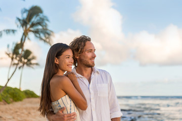 Obraz premium Relaxing casual couple watching sunset together on Waiohai beach, Poipu in Kauai, Hawaii. Beautiful multiracial couple relaxing on summer holidays.