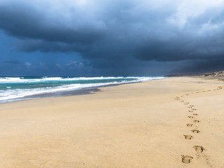 Endless, deserted, lonely sandy beach up to the horizon with footprints in the sand, small waves and a turquoise colored ocean at sunny weather.