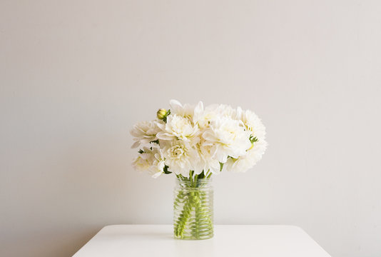 White dahlias in glass jar on white table against neutral background