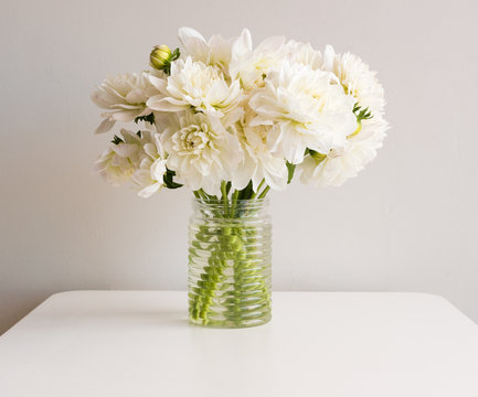 White dahlias in glass jar on white table against neutral background