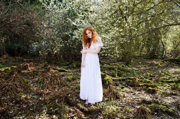 Beautiful young woman with cascading long red hair, wearing a long white dress, posing in a...