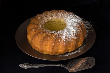 Homemade vanilla bundt cake with lemon, sprinkled with powdered sugar icing on a brown plate on a black background. Selective focus