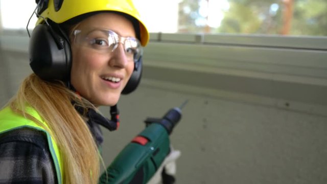 Young Female construction worker drilling concrete wall with the drill and smiling at the camera.